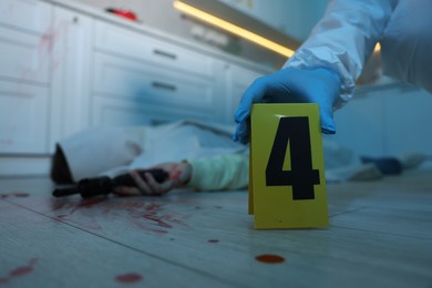 Photo of Forensic expert with numbered marker stand and dead body with gun at crime scene on floor indoors, selective focus