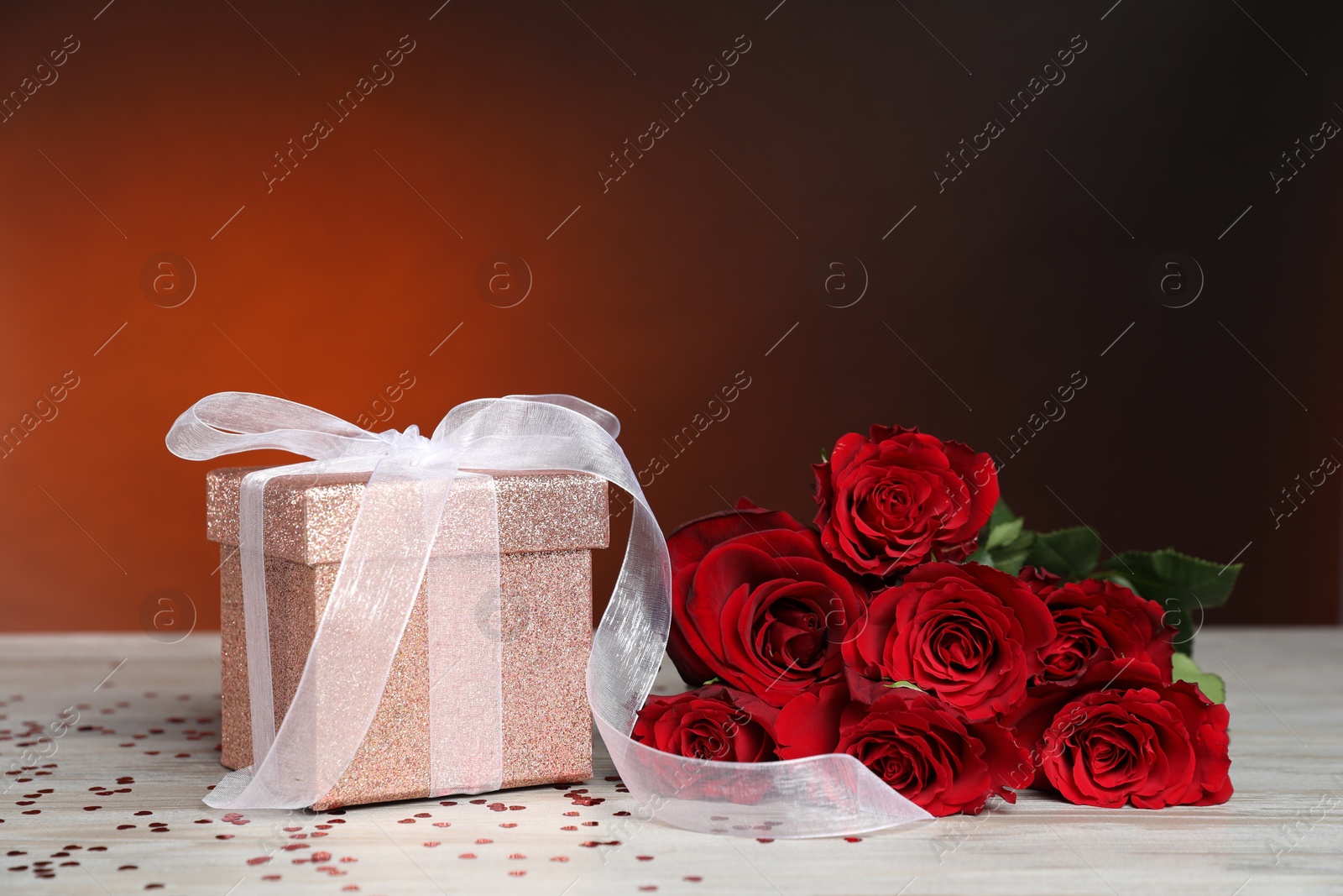 Photo of Beautiful gift box, roses and confetti on white wooden table against red background, closeup. Romantic present