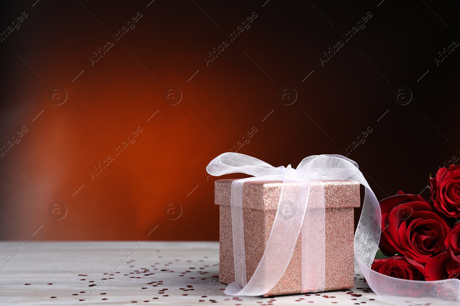 Photo of Beautiful gift box, roses and confetti on white wooden table against red background, closeup with space for text. Romantic present