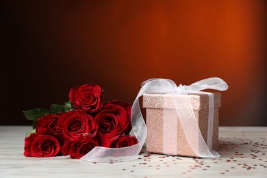 Beautiful gift box, roses and confetti on white wooden table against red background, closeup. Romantic present