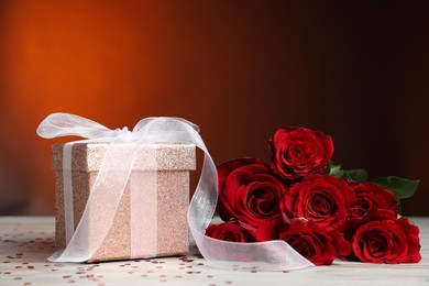 Photo of Beautiful gift box, roses and confetti on white wooden table against red background, closeup. Romantic present