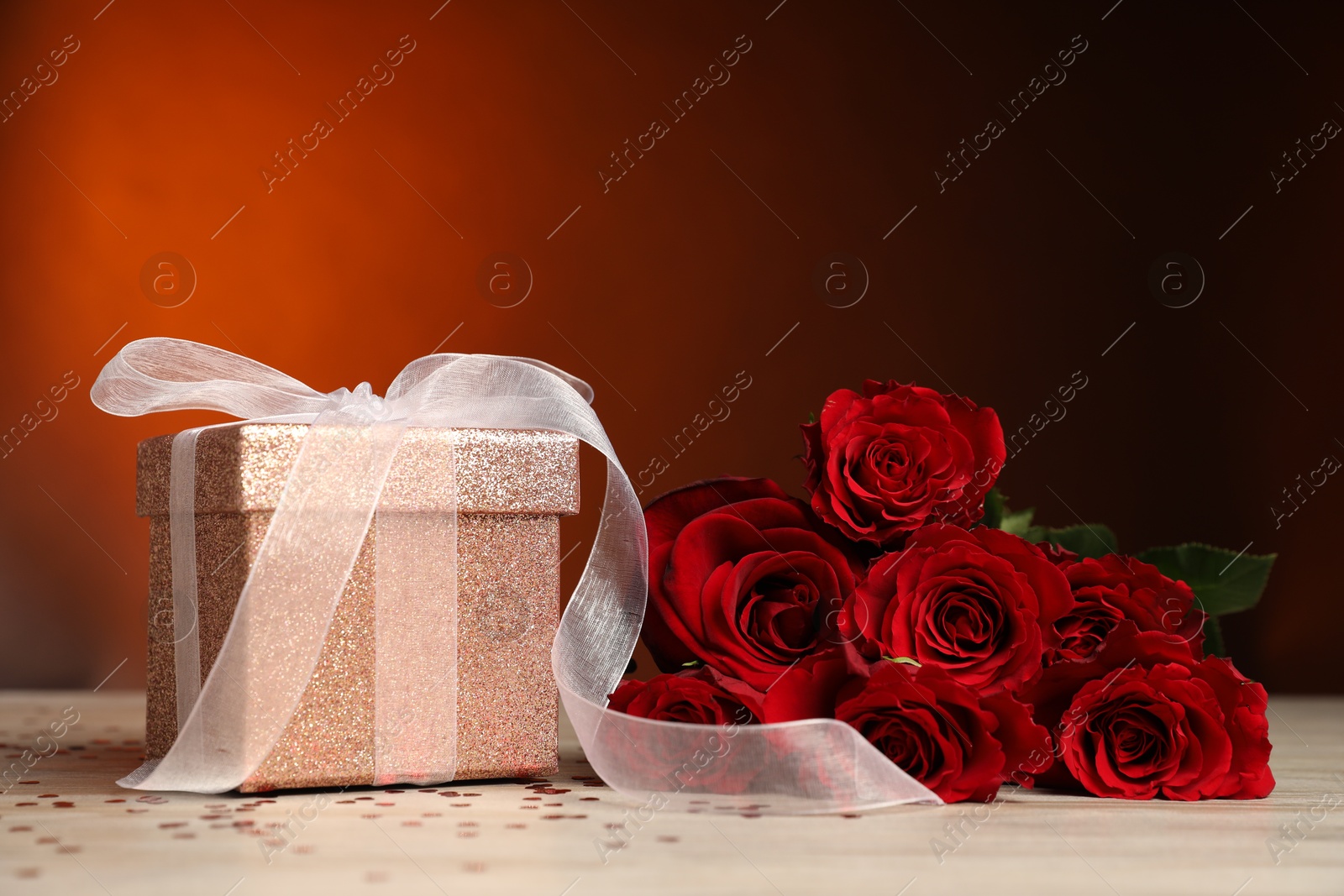 Photo of Beautiful gift box, roses and confetti on white wooden table against red background, closeup. Romantic present