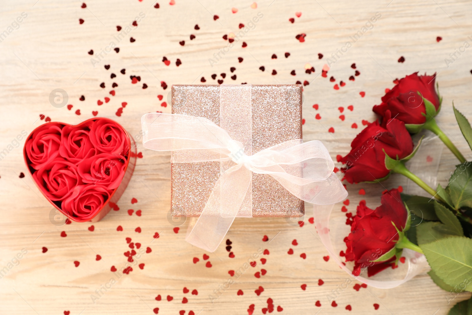 Photo of Romantic gift, roses and confetti in shape of hearts on light wooden table, flat lay. Happy Valentine's day