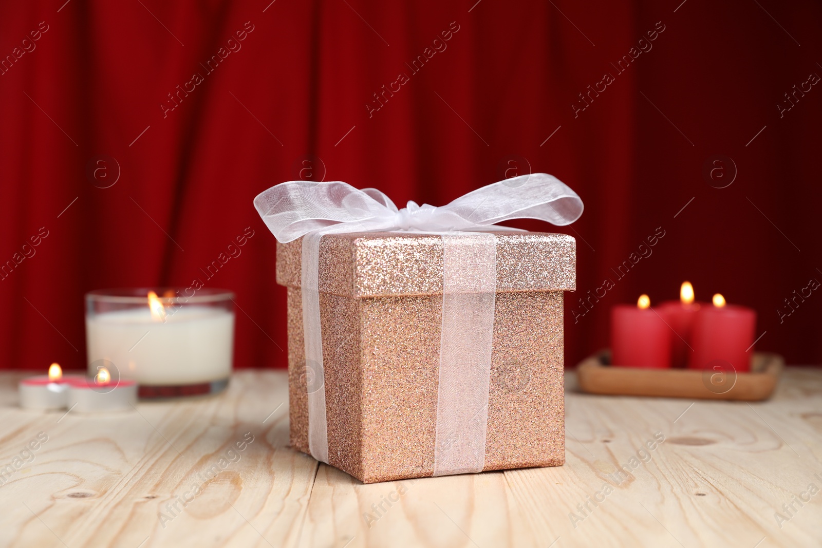 Photo of Beautiful gift box and burning candles on wooden table against red curtain. Romantic present