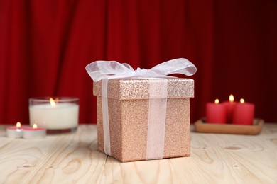 Photo of Beautiful gift box and burning candles on wooden table against red curtain. Romantic present