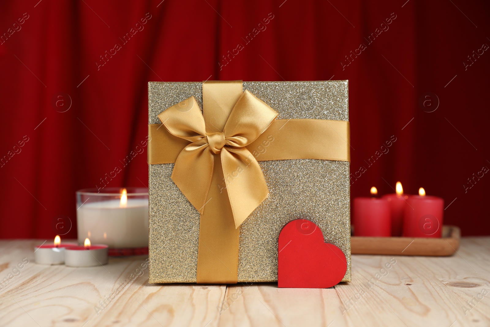 Photo of Beautiful gift box, decorative heart and burning candles on wooden table against red curtain. Romantic present