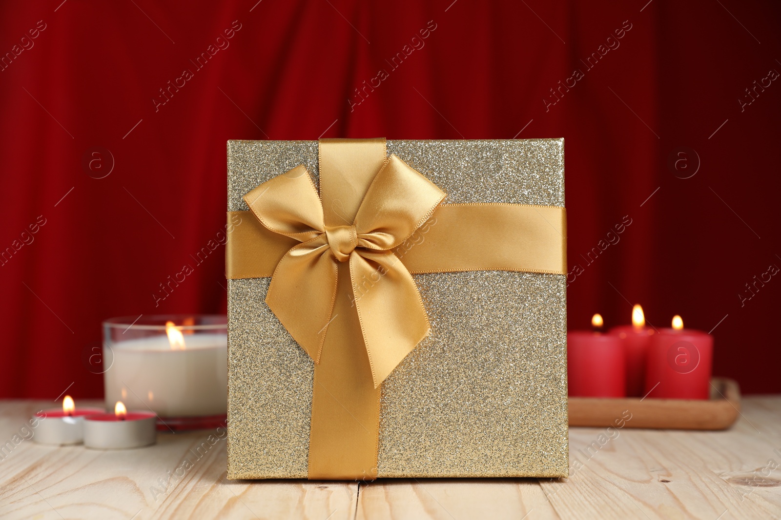 Photo of Beautiful gift box and burning candles on wooden table against red curtain. Romantic present