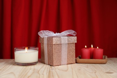 Beautiful gift box and burning candles on wooden table against red curtain. Romantic present