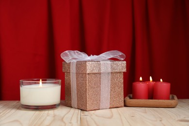 Photo of Beautiful gift box and burning candles on wooden table against red curtain. Romantic present