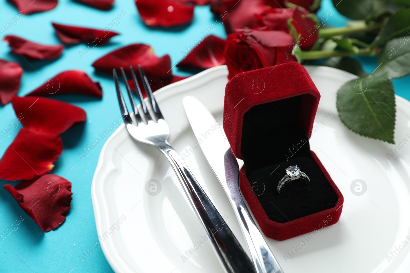 Photo of Beautiful setting for romantic dinner, engagement ring and rose petals on light blue background, closeup