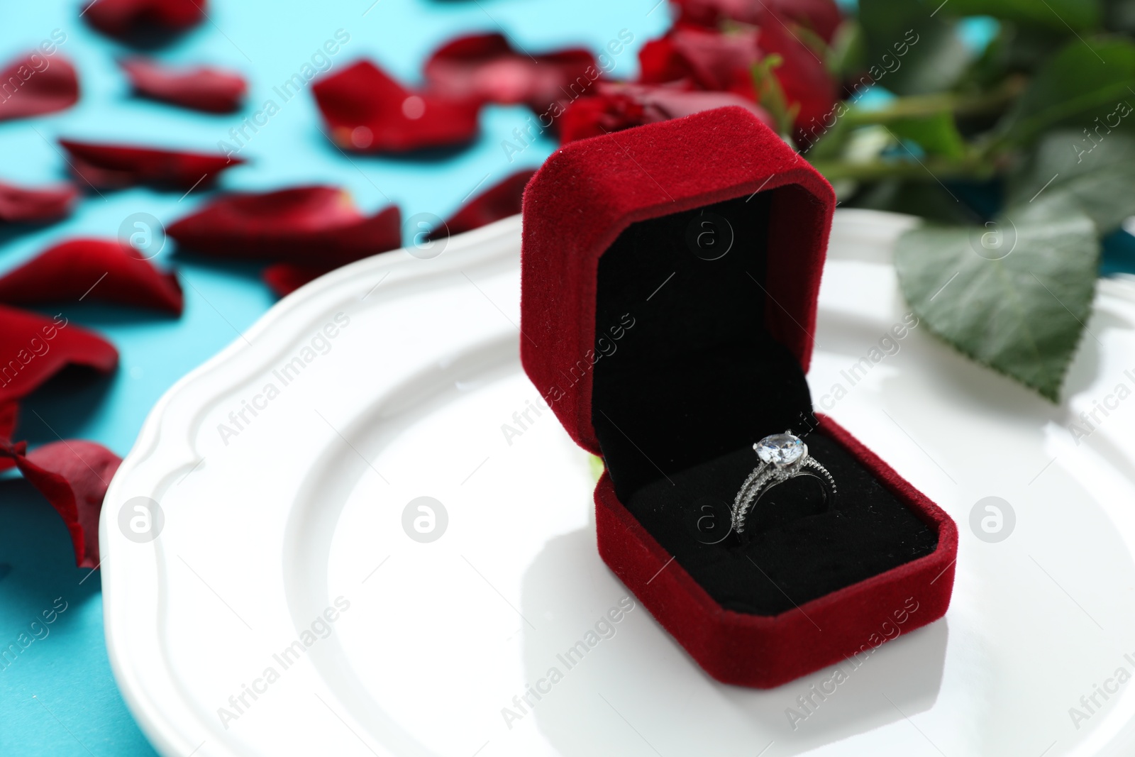 Photo of Beautiful setting for romantic dinner, engagement ring and rose petals on light blue background, closeup