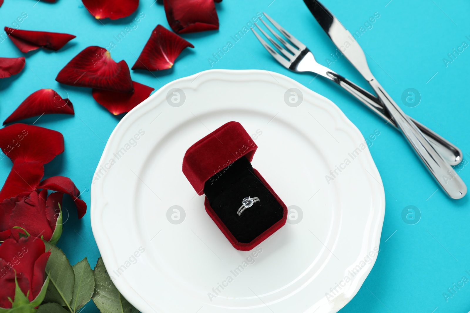 Photo of Beautiful setting for romantic dinner, engagement ring and rose petals on light blue background, flat lay