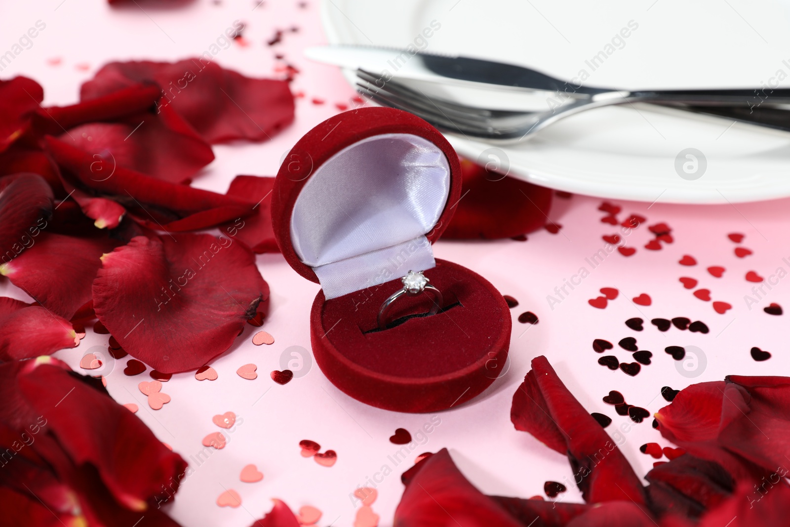 Photo of Beautiful setting for romantic dinner, engagement ring and rose petals on light pink table, closeup