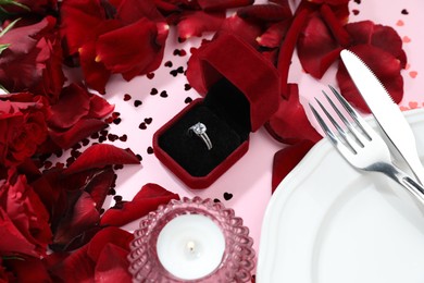 Photo of Beautiful setting for romantic dinner, engagement ring and rose petals on light pink table, closeup