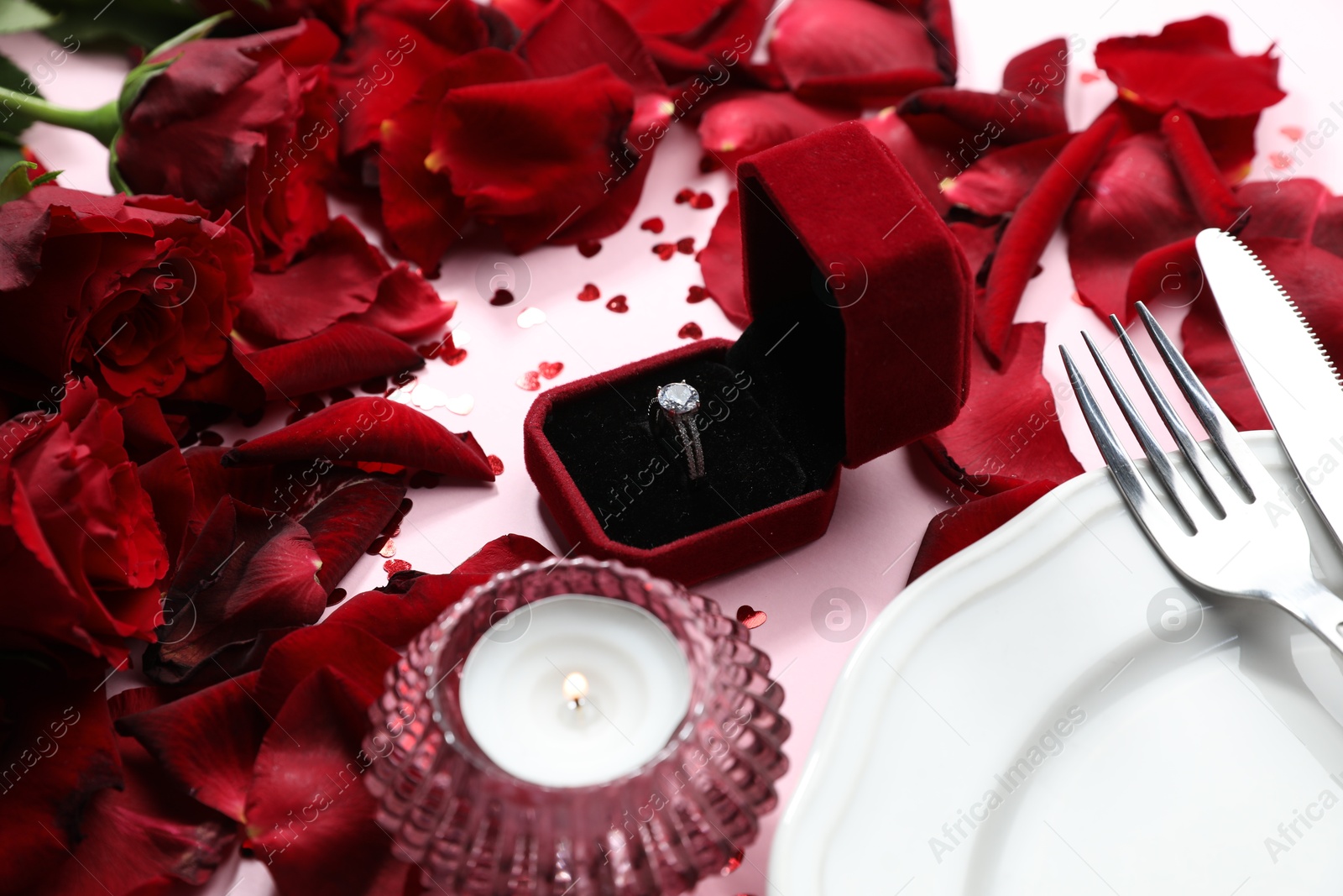 Photo of Beautiful setting for romantic dinner, engagement ring and rose petals on light pink table, closeup