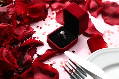 Photo of Beautiful setting for romantic dinner, engagement ring and rose petals on light pink table, closeup