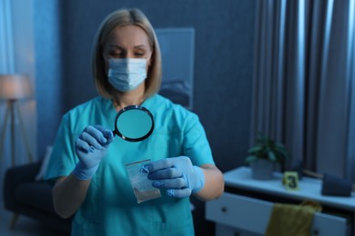 Forensic expert examining plastic bag with drug powder through magnifying glass at crime scene indoors, selective focus. Space for text