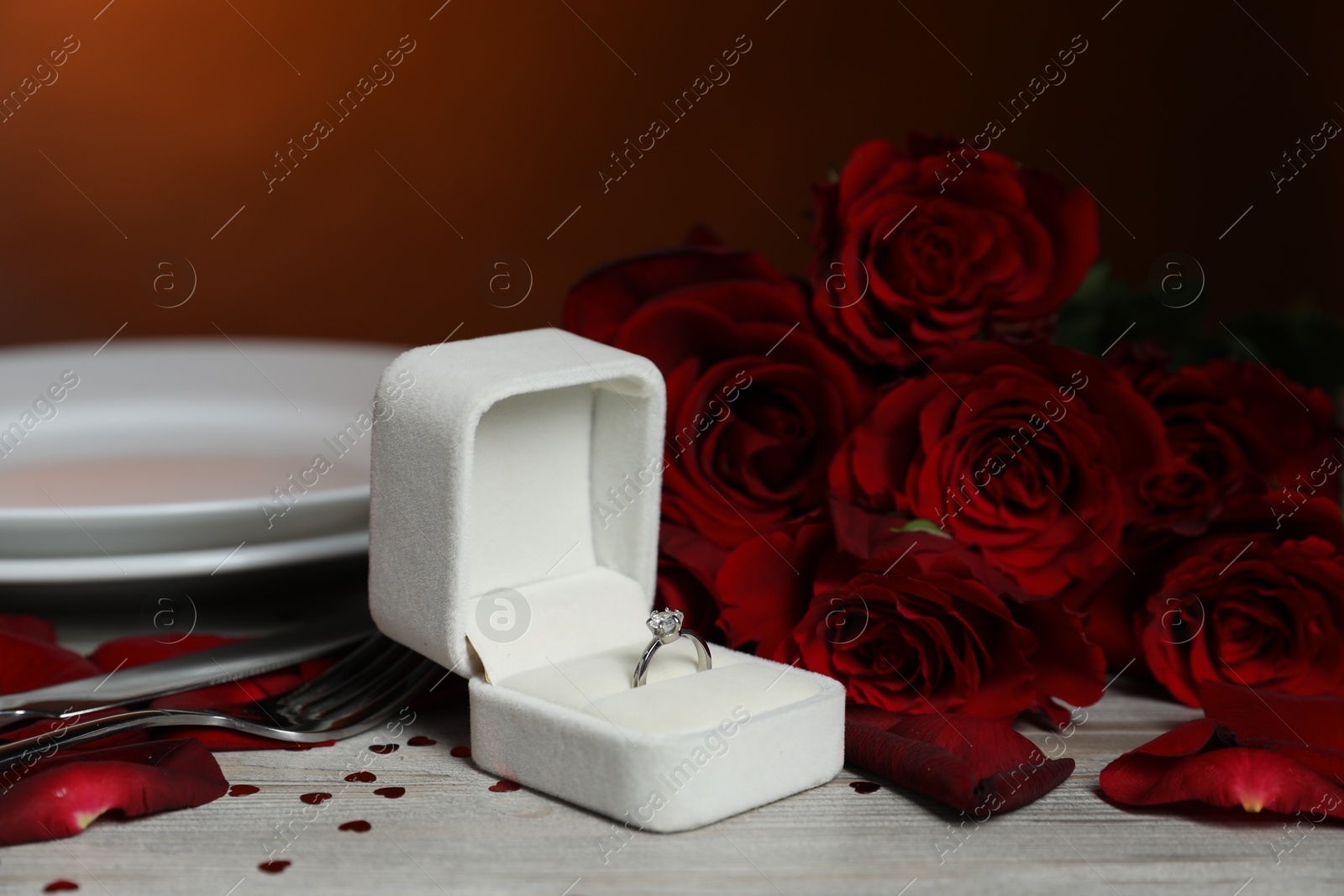 Photo of Engagement ring in box, roses, confetti and tableware on light wooden table against dark background, closeup. Romantic dinner