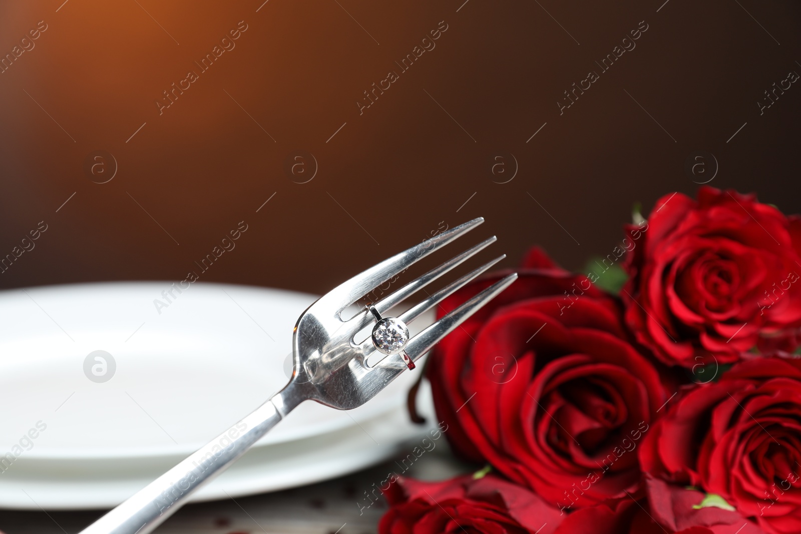 Photo of Fork with engagement ring, roses and plate against dark background, closeup. Romantic dinner