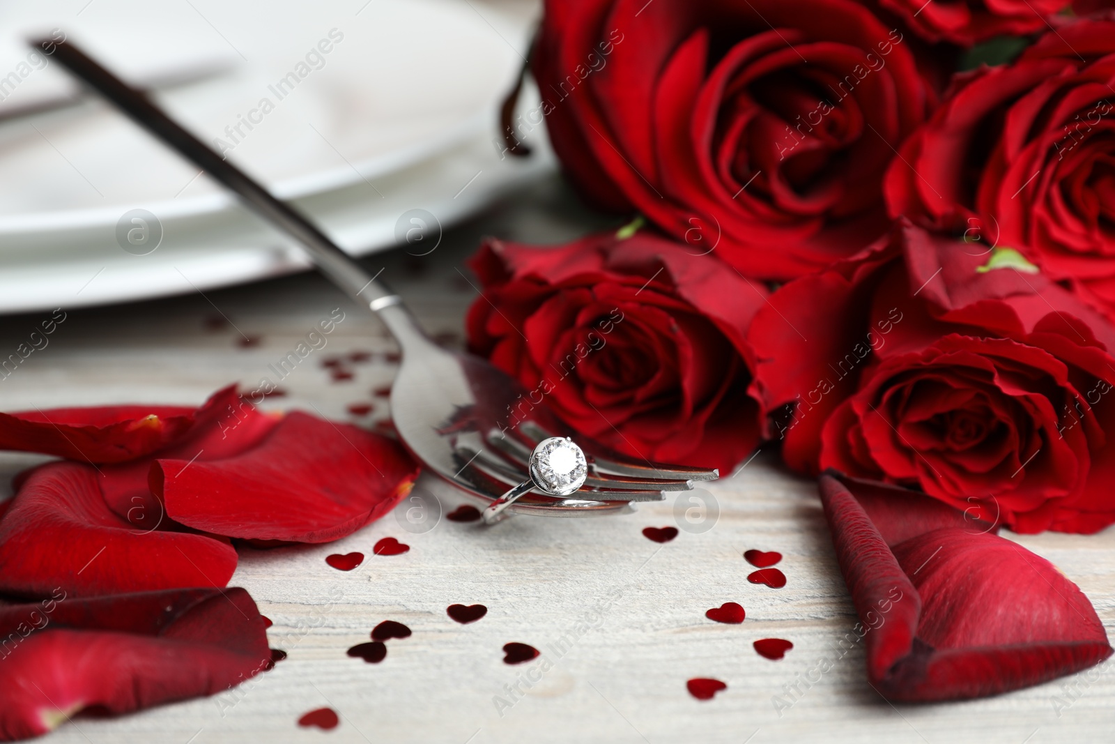 Photo of Fork with engagement ring, roses and confetti on light wooden table, closeup. Romantic dinner