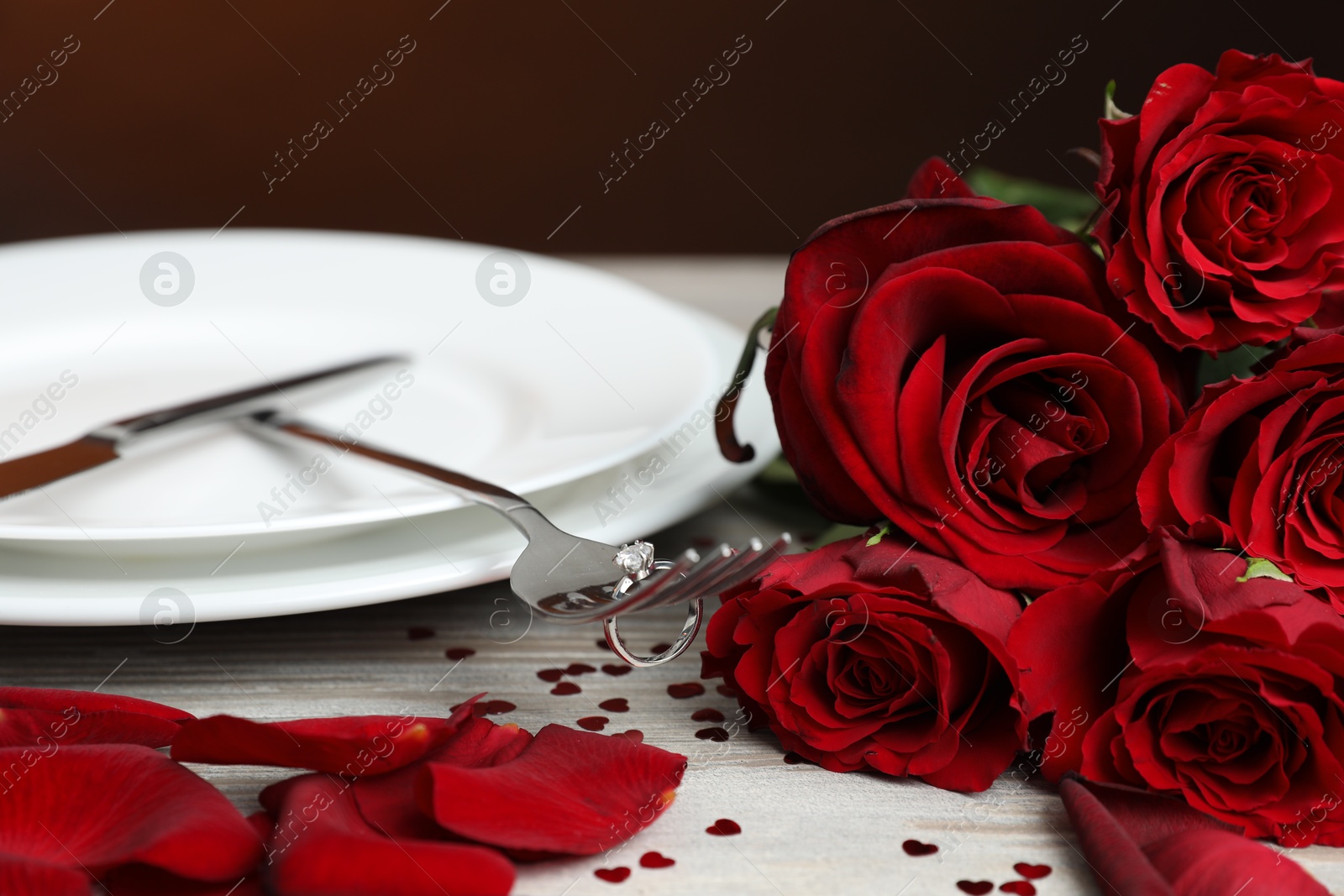 Photo of Fork with engagement ring, plates, roses and confetti on light wooden table, closeup. Romantic dinner