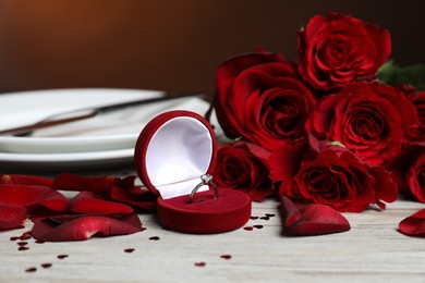 Photo of Engagement ring in box, roses, confetti and tableware on light wooden table, closeup. Romantic dinner