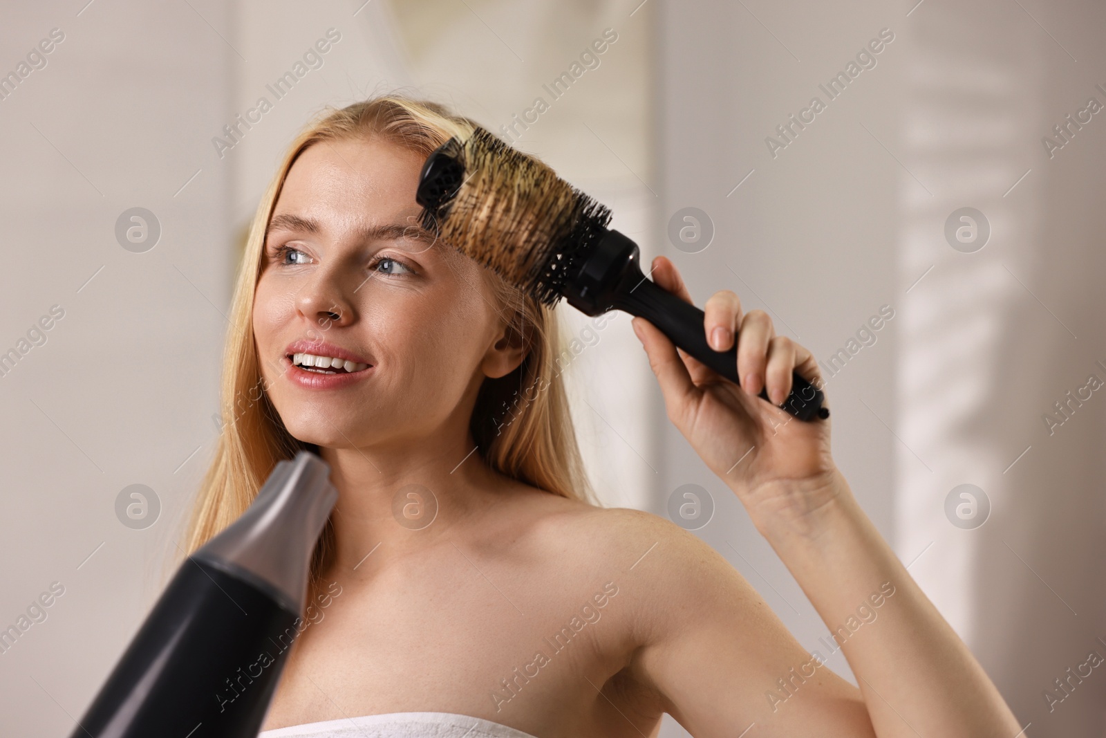 Photo of Beautiful young woman styling her hair with hairdryer and brush in bathroom