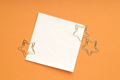 Photo of Star shaped clips and blank paper note on orange background, flat lay