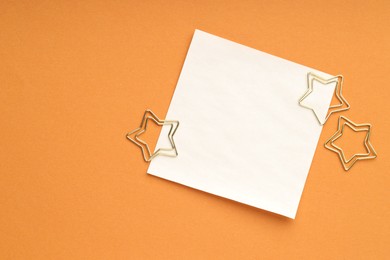 Photo of Star shaped clips and blank paper note on orange background, flat lay. Space for text