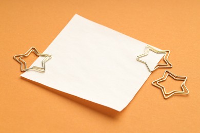 Photo of Star shaped clips and blank paper note on orange background, closeup