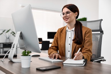 Photo of Woman taking notes while working on computer at table in office