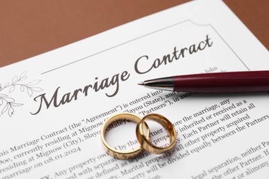 Photo of Marriage contract, pen and golden rings on brown background, closeup