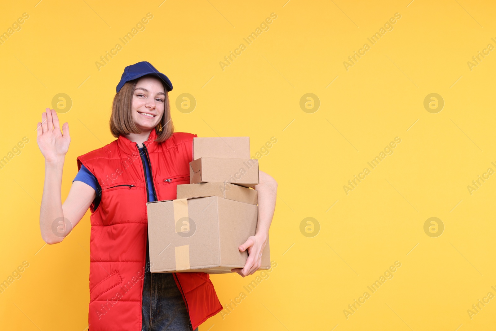 Photo of Happy postwoman with parcels waving hello on yellow background. Space for text