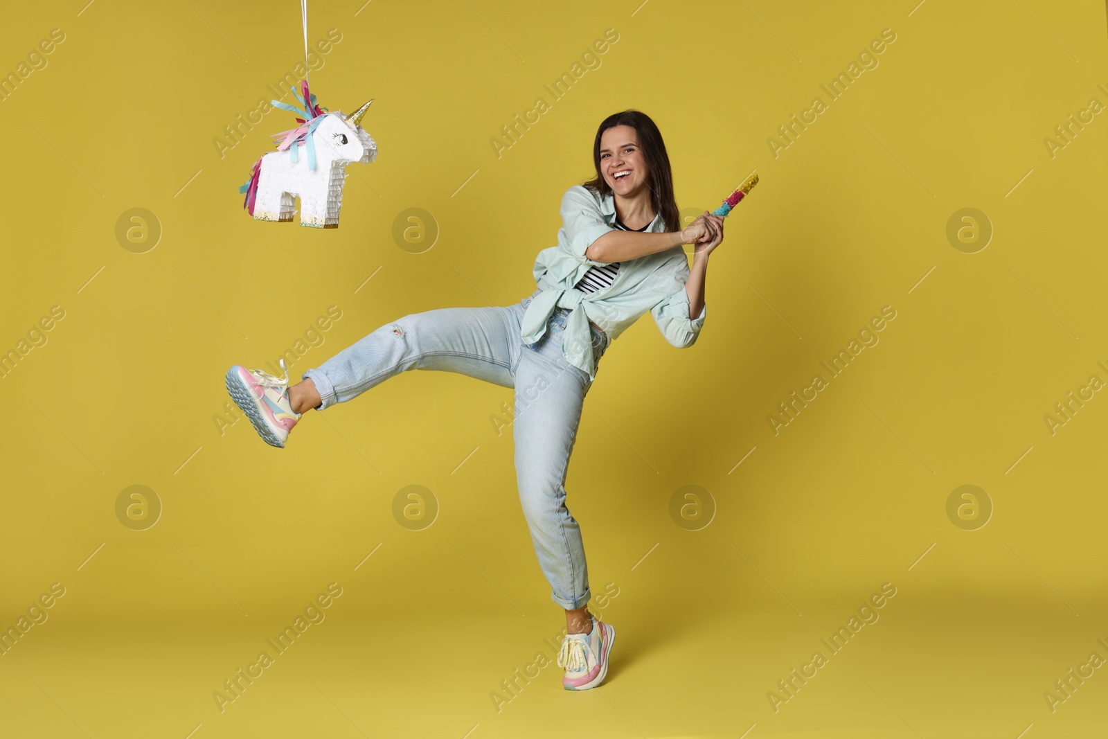 Photo of Happy woman hitting unicorn shaped pinata with stick on yellow background