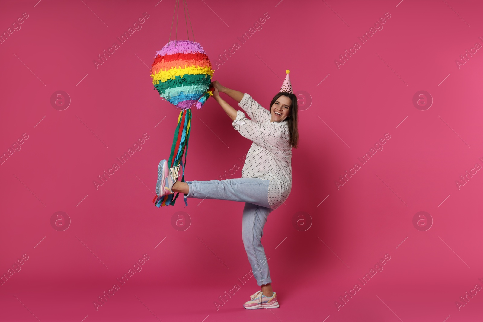 Photo of Happy woman hitting colorful pinata with stick on pink background