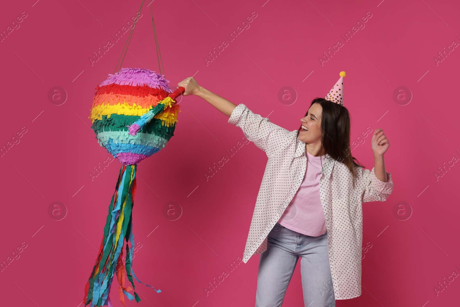 Photo of Happy woman hitting colorful pinata with stick on pink background