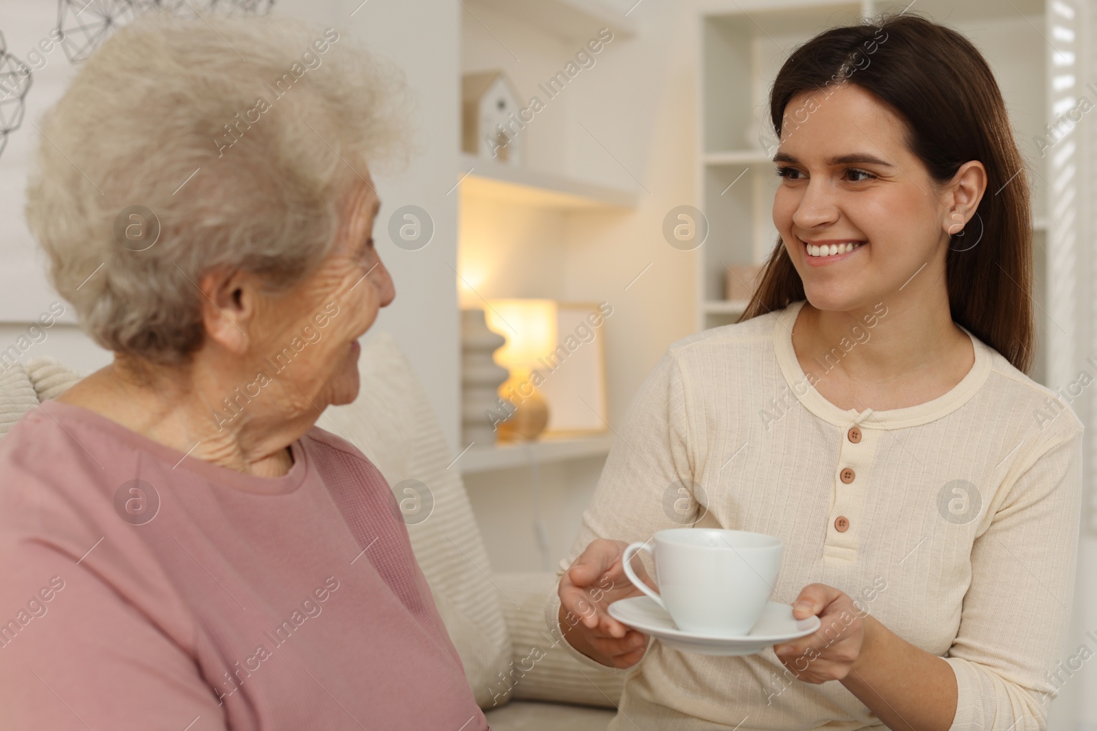 Photo of Granddaughter giving hot drink to her grandmother at home. Elderly care