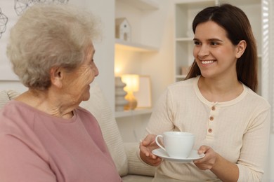 Photo of Granddaughter giving hot drink to her grandmother at home. Elderly care