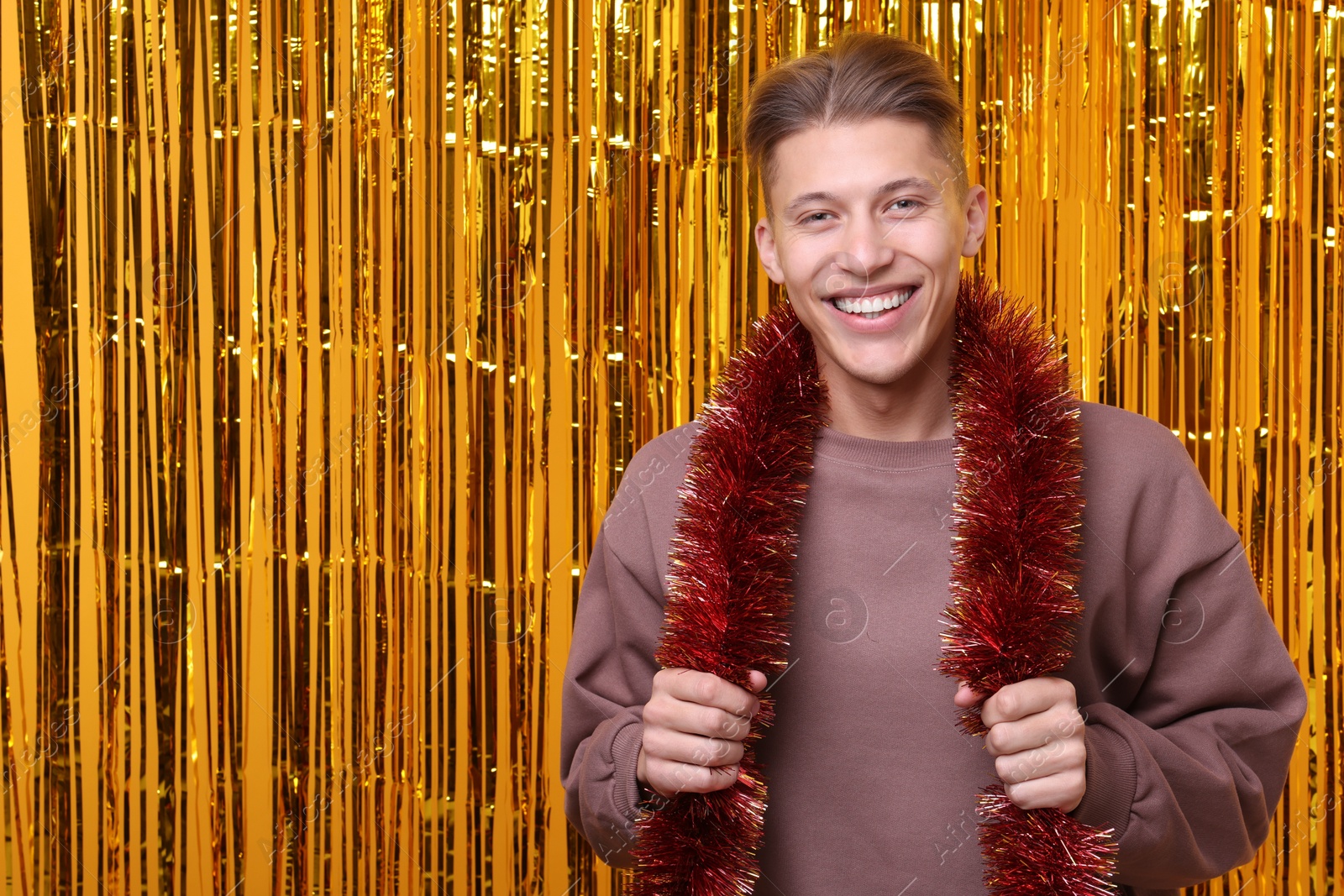 Photo of Happy young man with bright tinsel against golden foil curtain