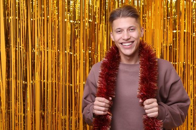 Photo of Happy young man with bright tinsel against golden foil curtain