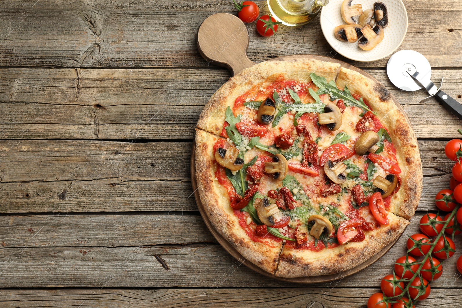 Photo of Tasty pizza with mushrooms, tomatoes, arugula and cutter on wooden table, flat lay. Space for text