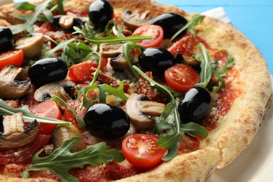 Tasty pizza with mushrooms, black olives and arugula on light blue table, closeup