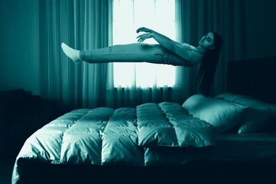 Image of Sleepwalking. Young woman floating above bed at home, toned in blue