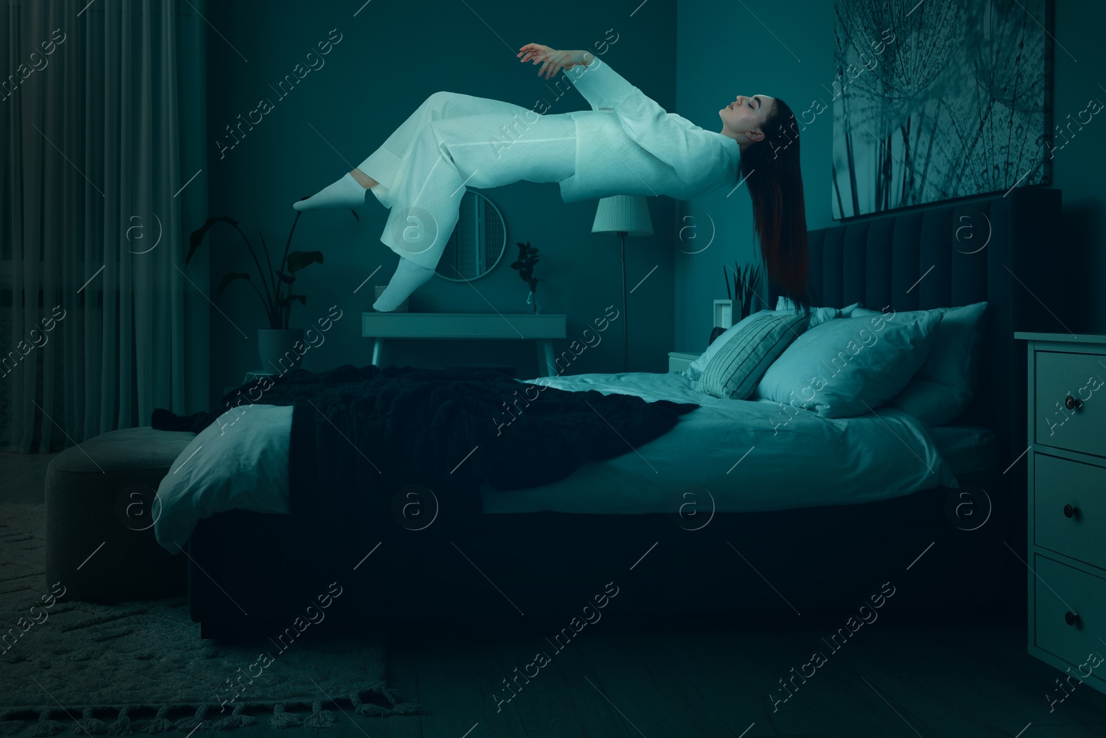 Image of Sleepwalking. Young woman floating over bed at home, toned in blue