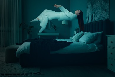 Image of Sleepwalking. Young woman floating over bed at home, toned in blue