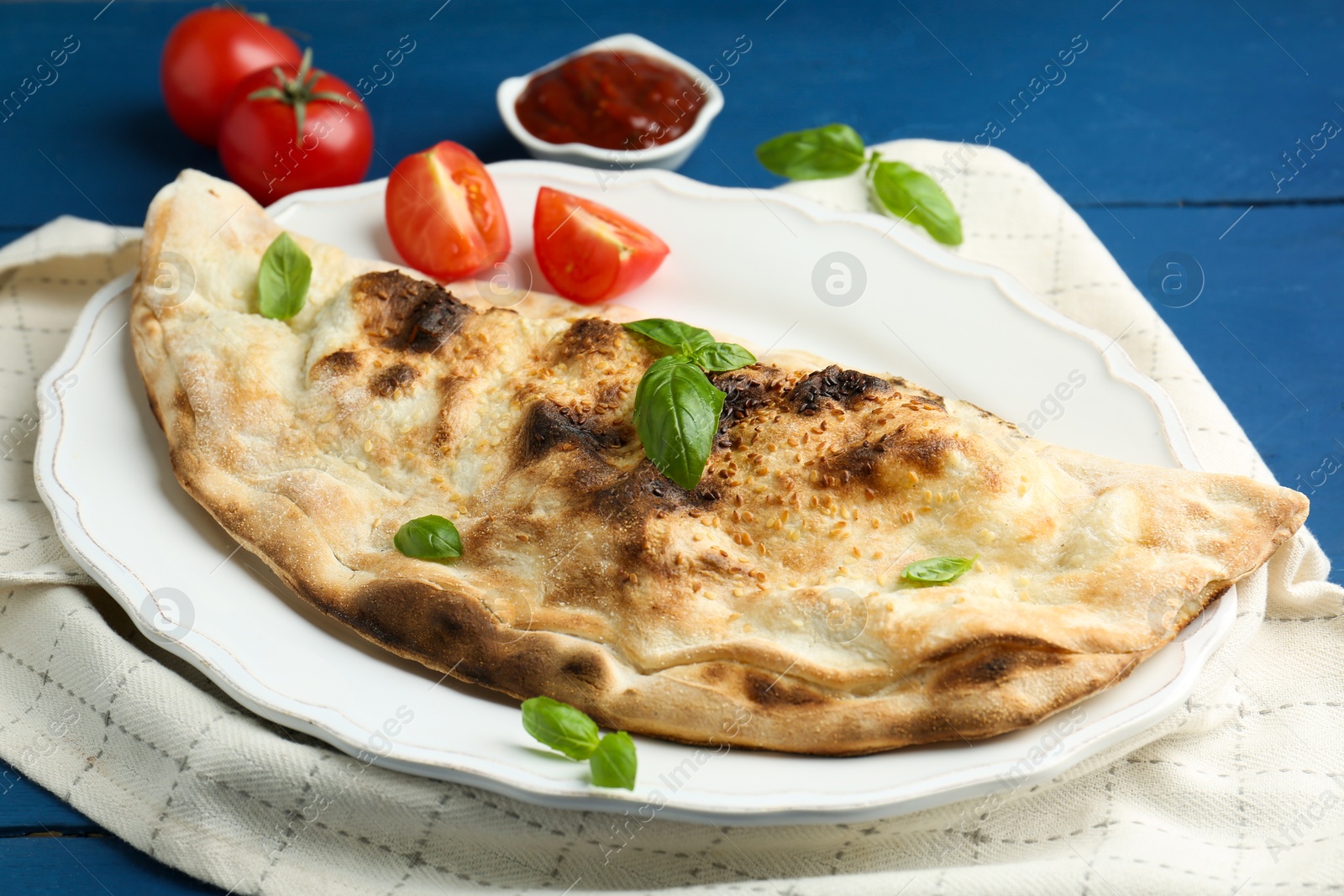 Photo of Tasty calzone with basil, tomatoes and sauce on blue wooden table, closeup