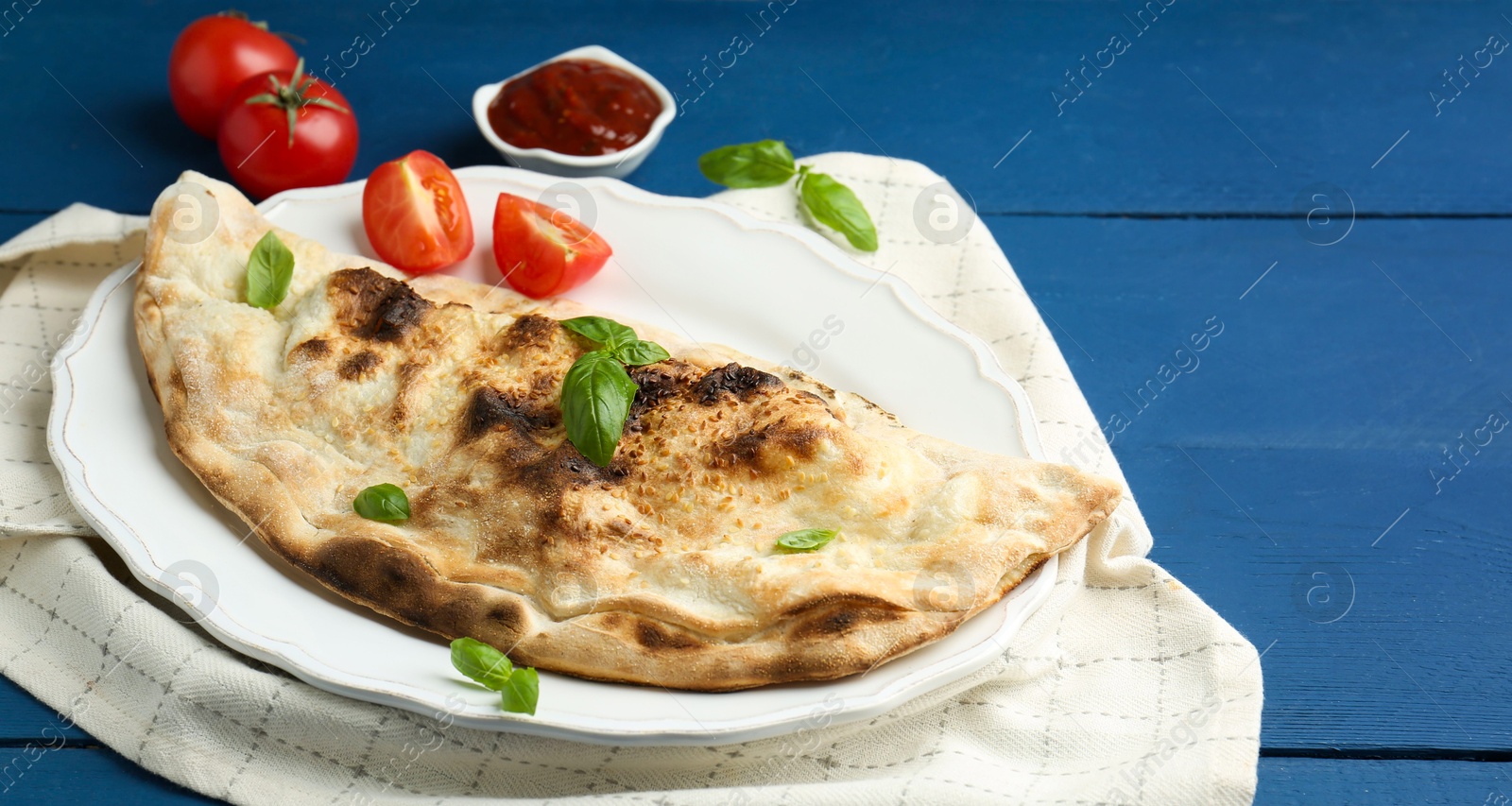 Photo of Tasty calzone with basil, tomatoes and sauce on blue wooden table, closeup