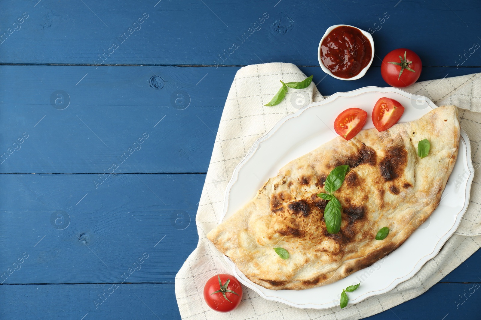 Photo of Tasty calzone with basil, tomatoes and sauce on blue wooden table, flat lay. Space for text