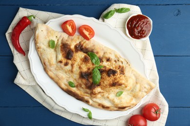 Photo of Tasty calzone with basil, tomatoes, chili pepper and sauce on blue wooden table, flat lay