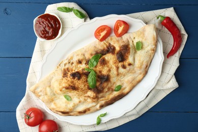 Photo of Tasty calzone with basil, tomatoes, chili pepper and sauce on blue wooden table, flat lay